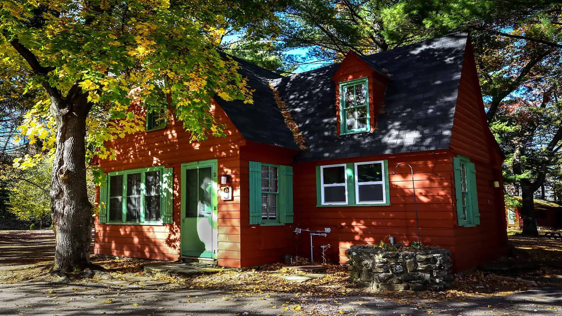 A quaint red cottage with green shutters is framed by vibrant autumn leaves.