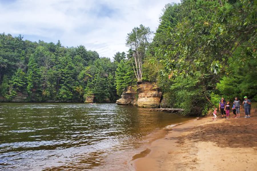 A sandy riverbank surrounded by lush greenery and several people walking along the shore.