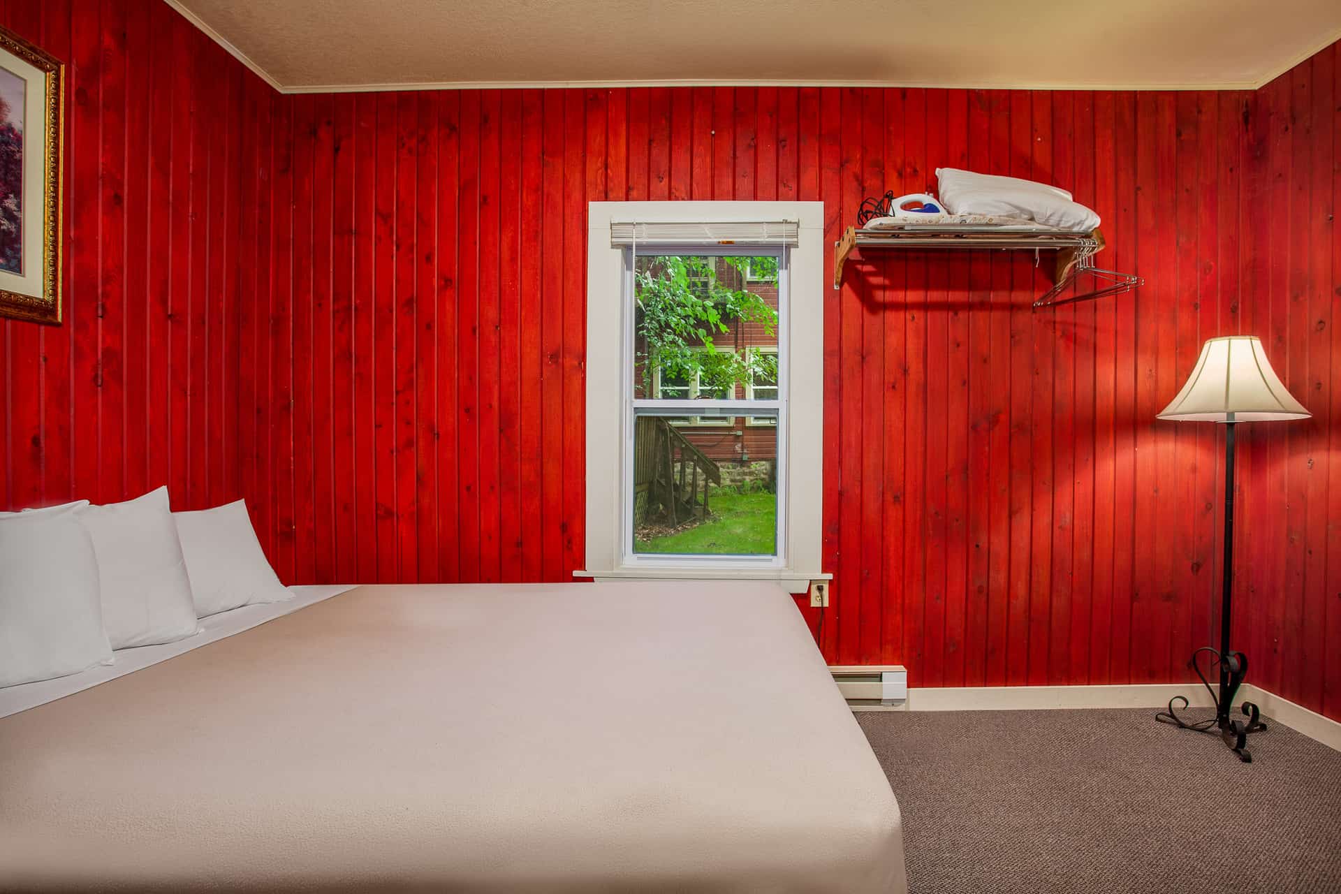 A cozy bedroom featuring bold red paneled walls, a white-covered bed, a classic floor lamp, and a wall-mounted shelf.