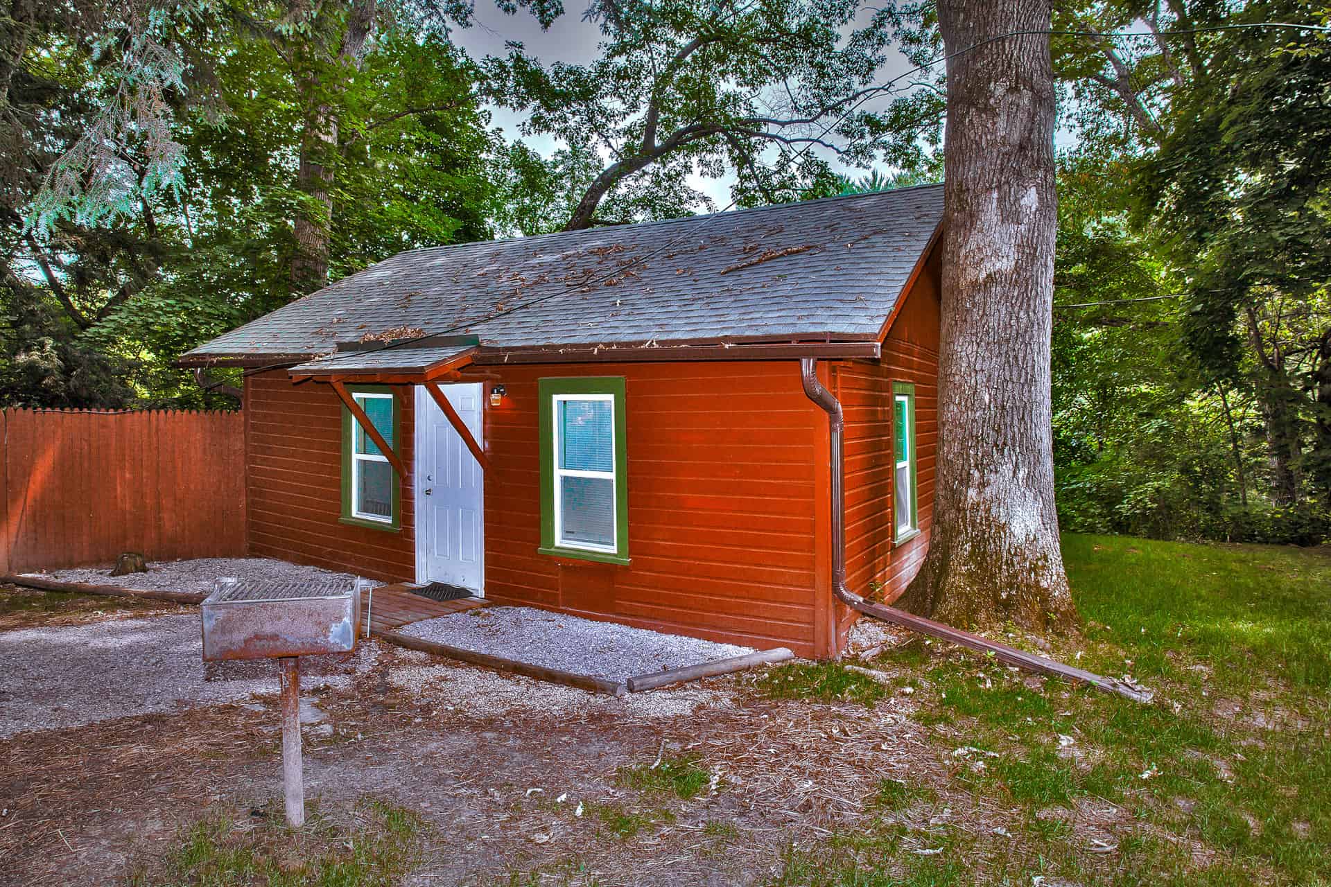 A charming rustic cabin with a red exterior and green-trimmed windows, nestled among trees, featuring a small porch and a nearby BBQ grill.