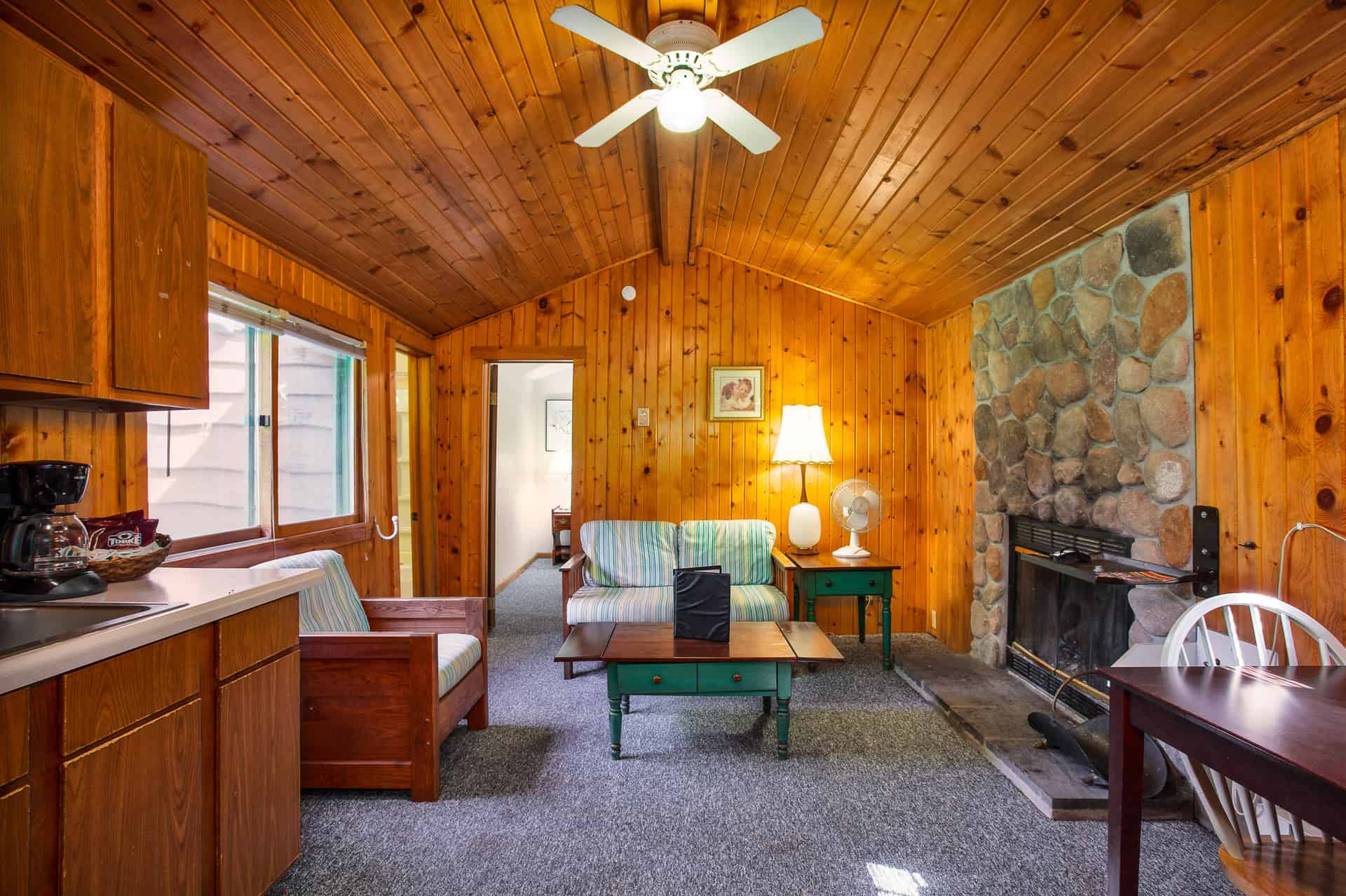 A cozy cabin interior with knotty pine walls, a stone fireplace, a living area with a sofa, and a kitchen counter with a coffee maker.