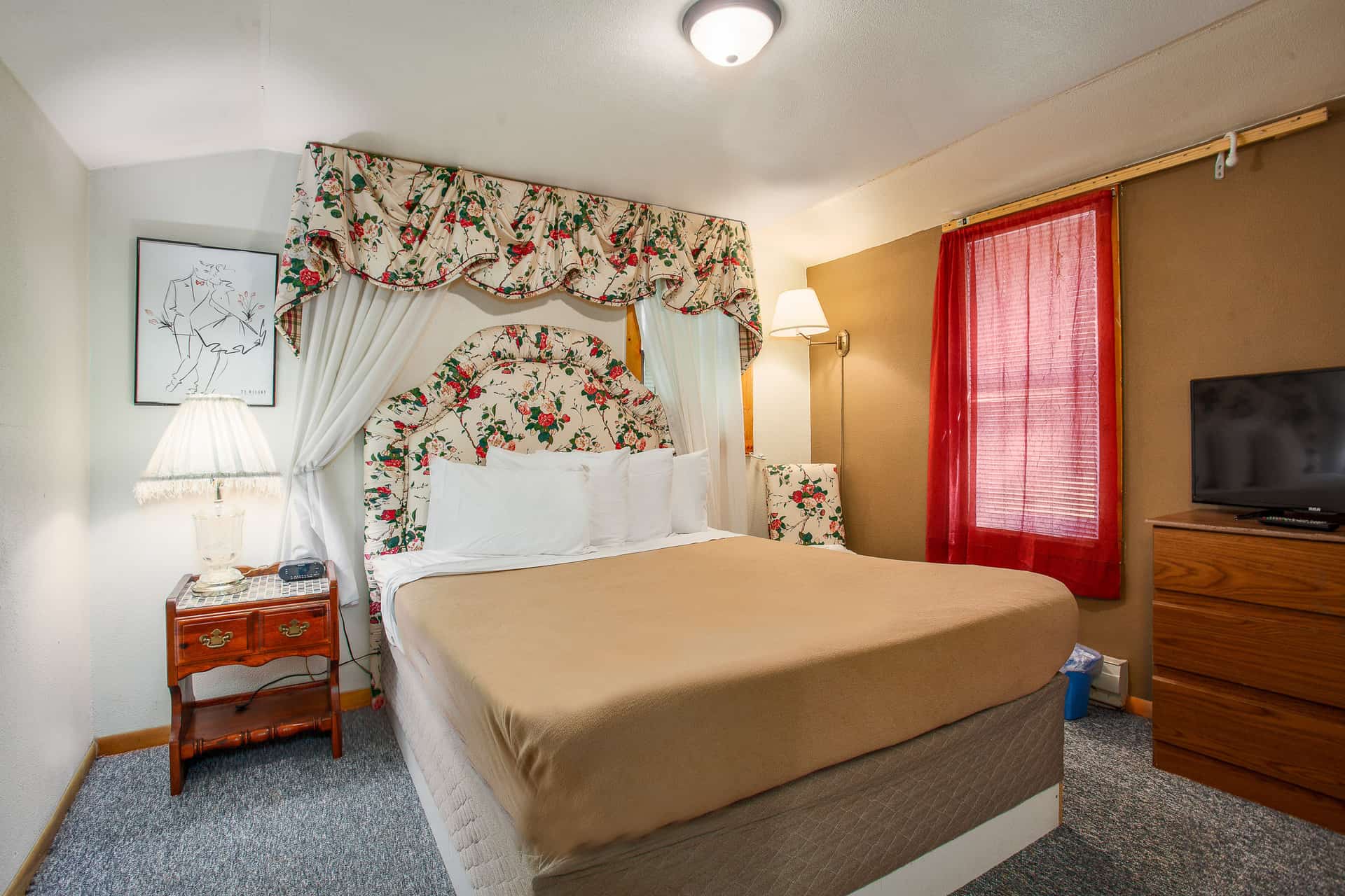 An inviting bedroom featuring a queen bed with a floral headboard and canopy, a red sheer curtain over a window, a dresser with a TV, and a nightstand.