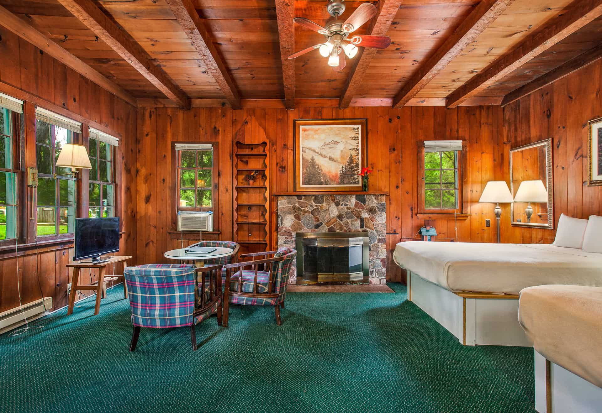 A cozy, rustic room with wood-paneled walls and ceiling, a stone fireplace, two full beds, a small seating area, and a television by the window.