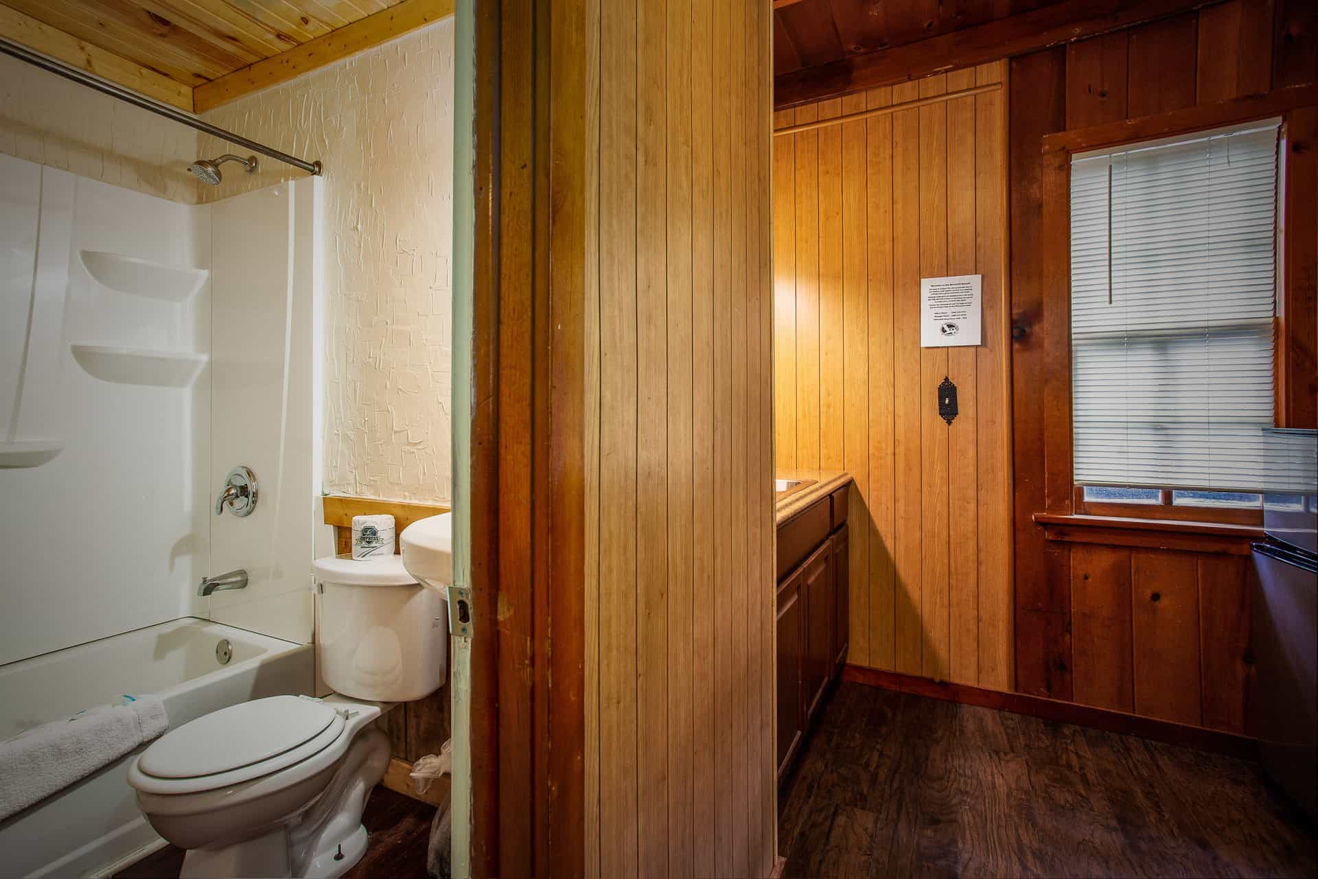 A split view of a rustic bathroom with wood-paneled walls and a window, leading into a separate area with a toilet and shower.