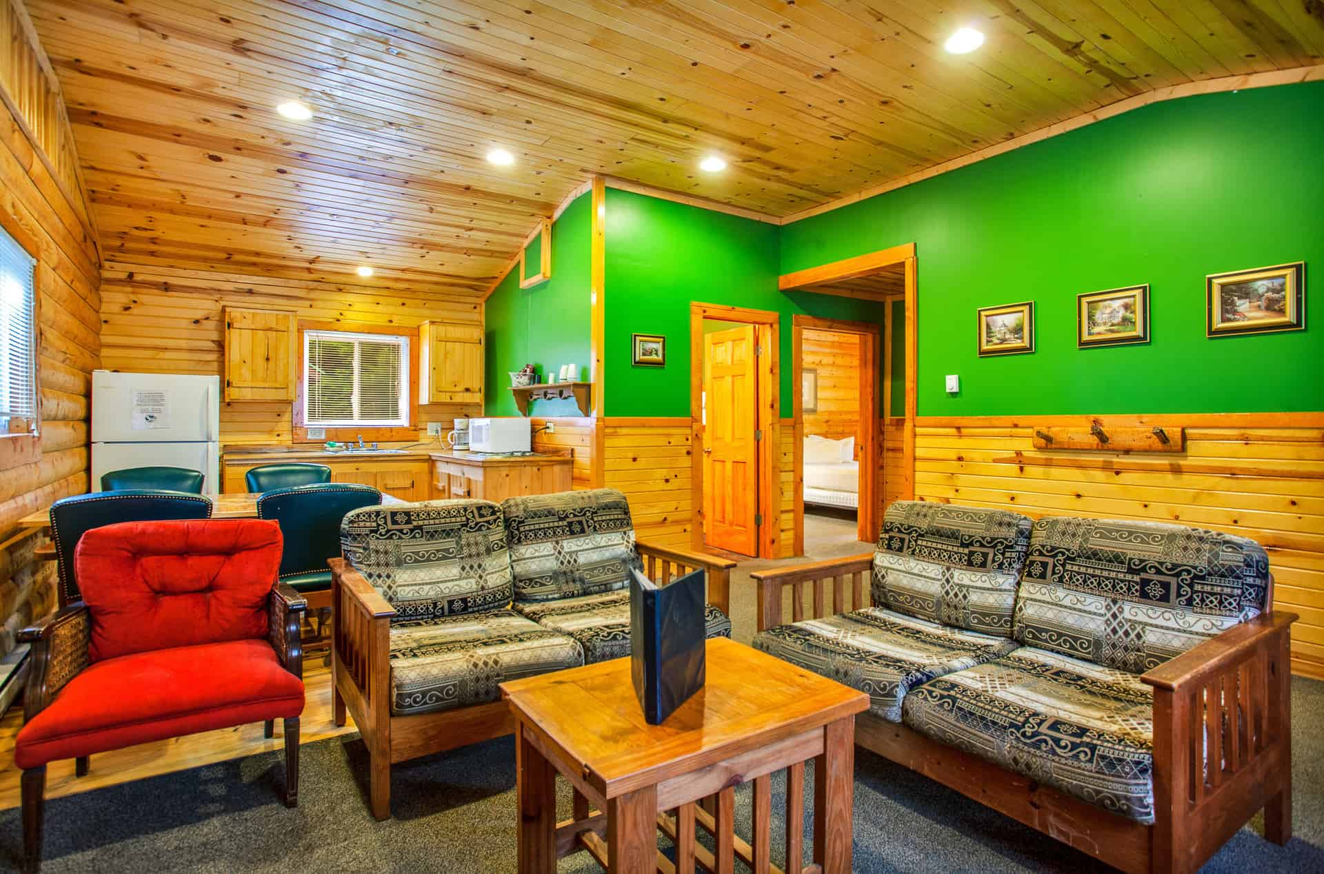 A vibrant cabin living area with wood paneling and green walls, featuring two sofas, a red armchair, a dining area, and a glimpse of a bedroom.