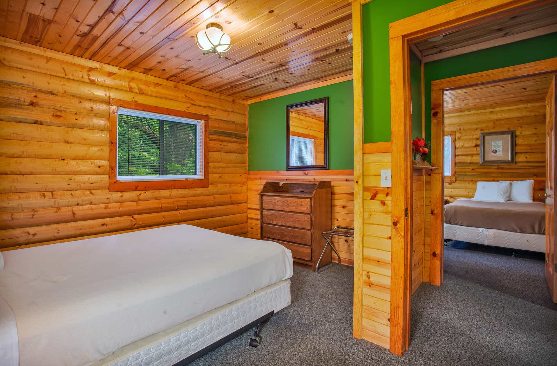 A warm cabin bedroom with wood-paneled and green walls, featuring a bed, dresser, and a doorway leading to another bedroom.