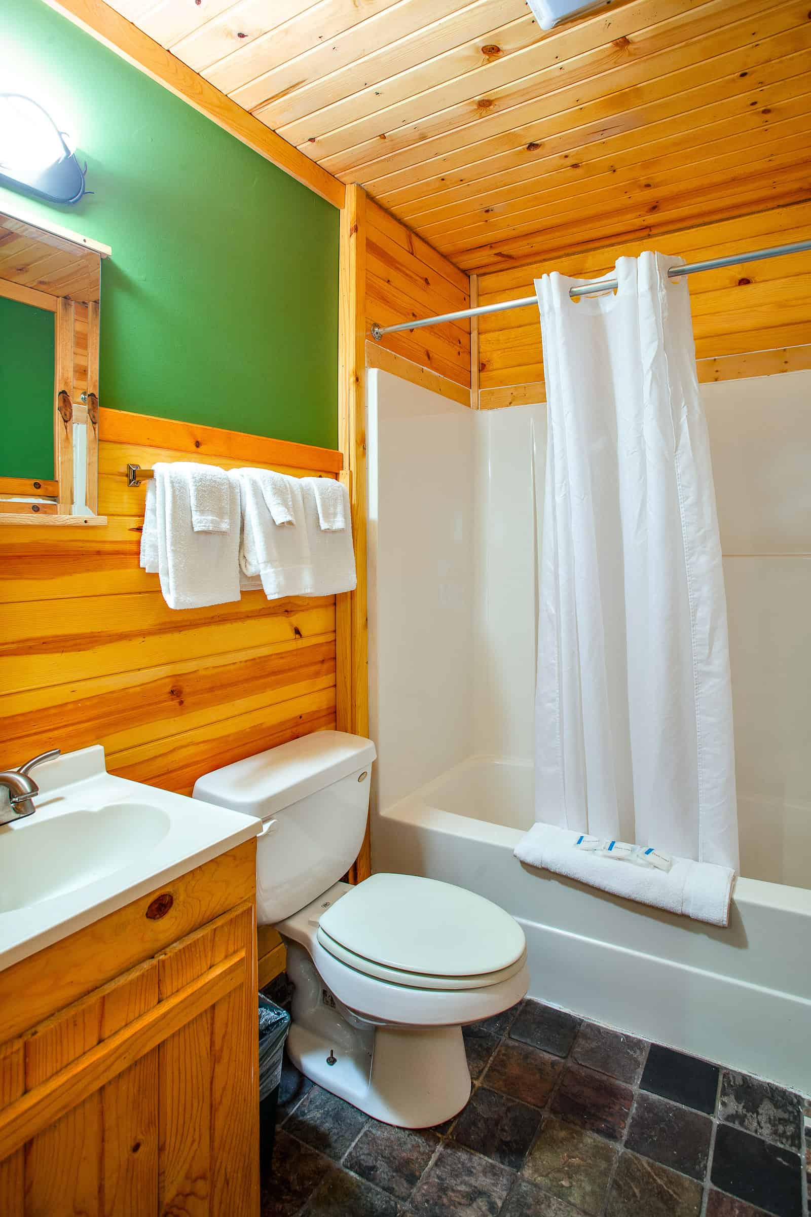 A rustic bathroom with warm wood paneling and green accent walls, featuring a toilet, sink vanity, and a shower/tub combo with a white curtain.