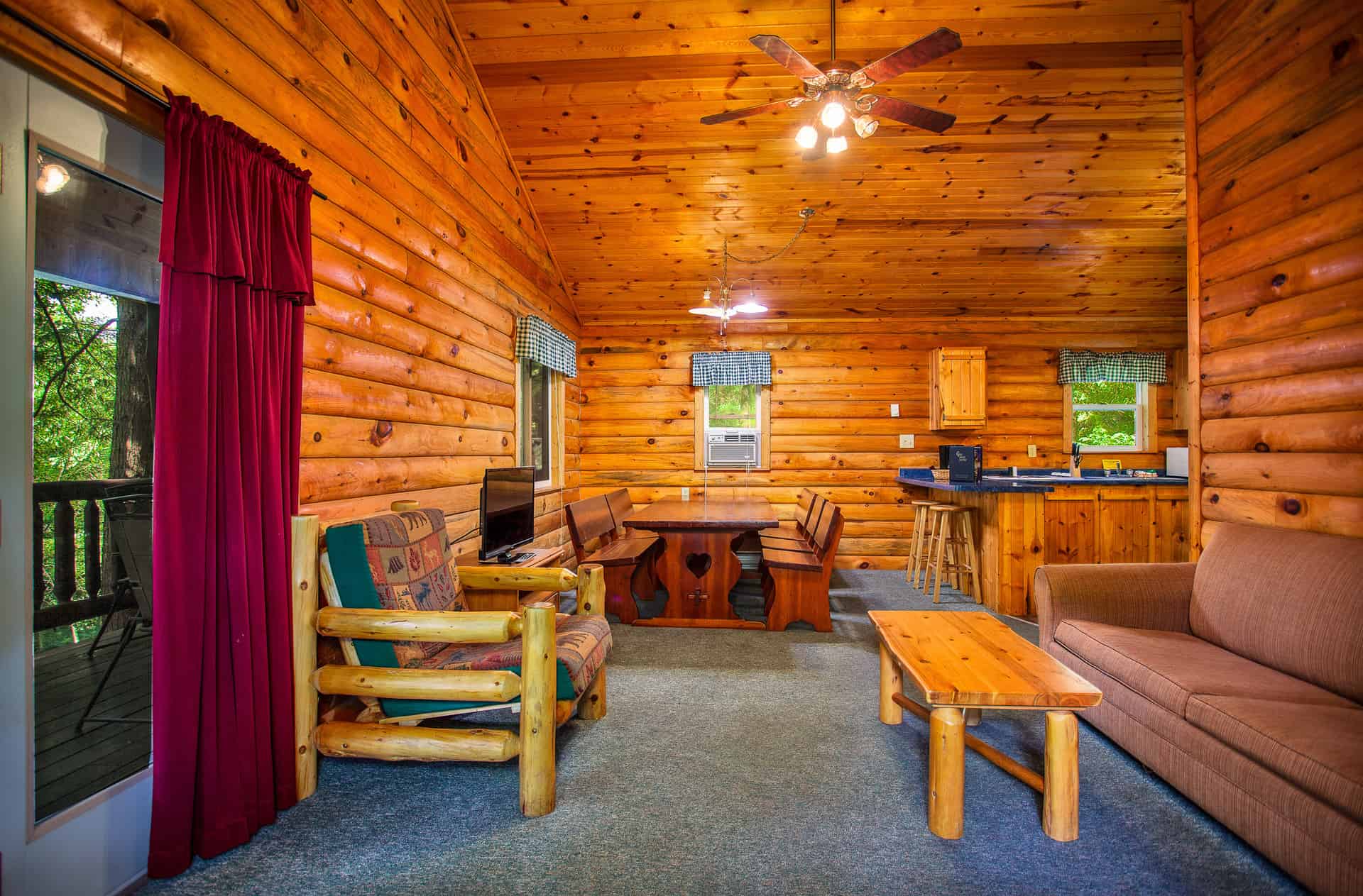 A spacious, rustic living area in a log cabin with wood-paneled walls and a vaulted ceiling, featuring two sofas, a coffee table, and a dining area.