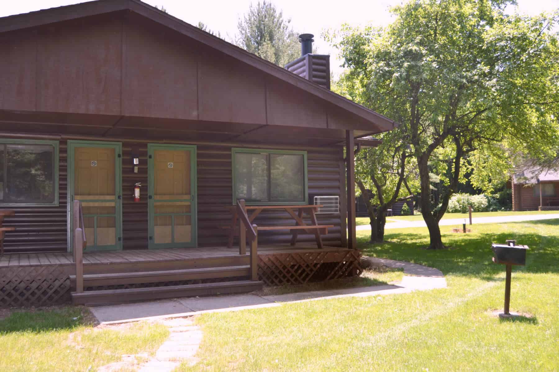 A cozy, rustic cabin with a wooden porch and a grill outside, surrounded by green grass and trees.