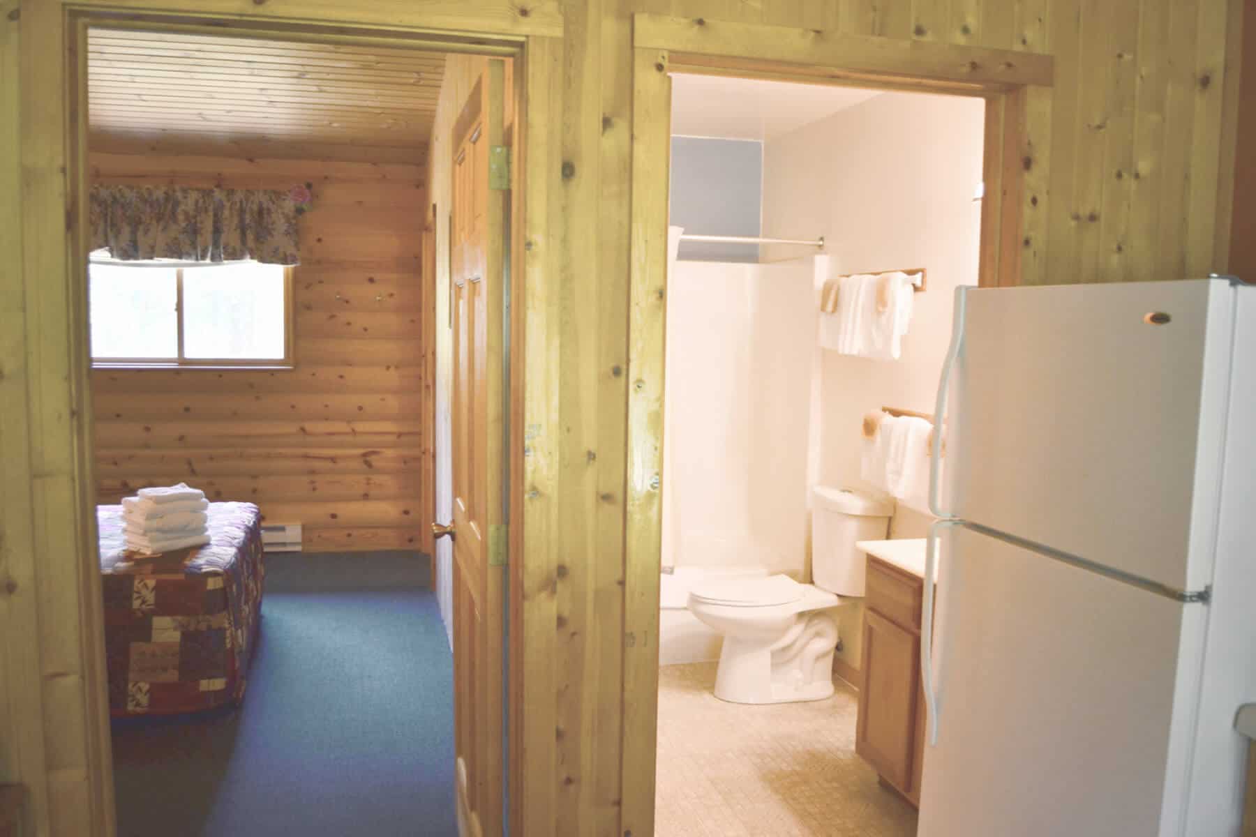 A practical cabin interior showcasing a bedroom with wood-paneled walls and a quilt-covered bed, adjacent to a bathroom and a kitchen.