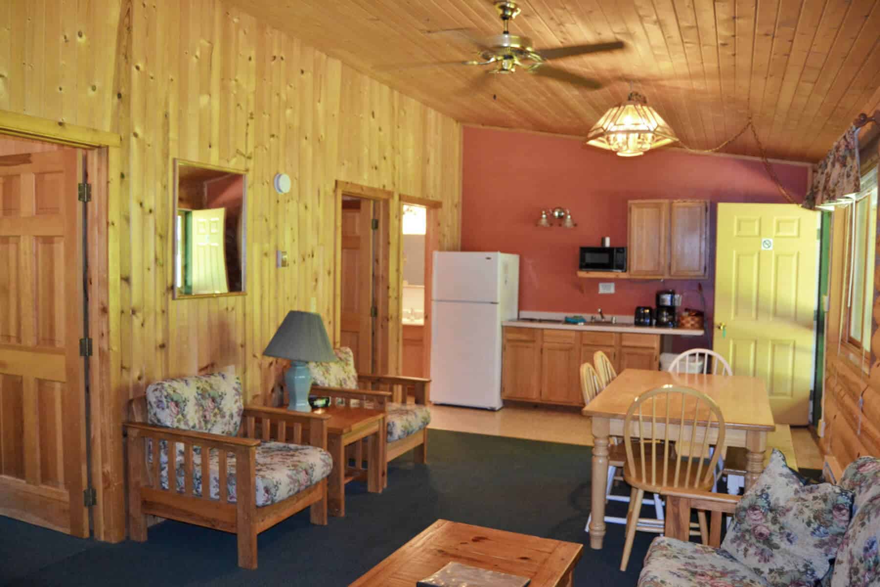 A charming cabin interior showcasing an open-plan living area with knotty pine walls, comfy seating, a dining table, and a compact kitchen.