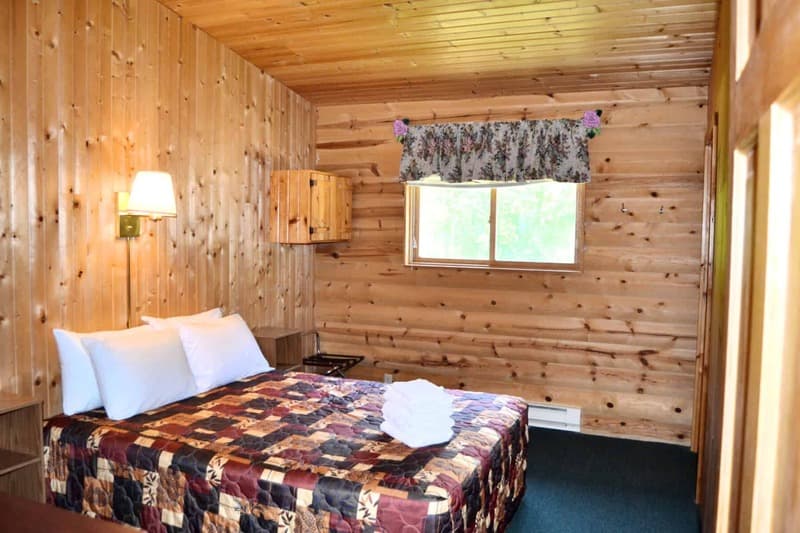 A rustic cabin bedroom featuring a queen bed with a patterned quilt, warm wood-paneled walls, a wall-mounted lamp, and a window with a valance.