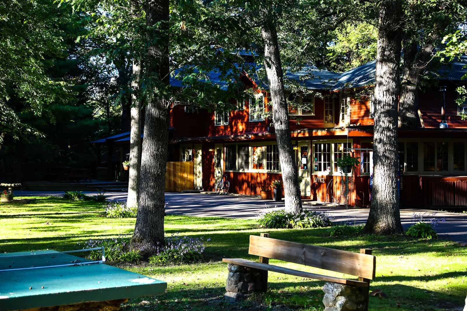 A red lodge building with numerous windows, nestled among tall trees, with a grassy foreground featuring picnic tables and a ping pong table.