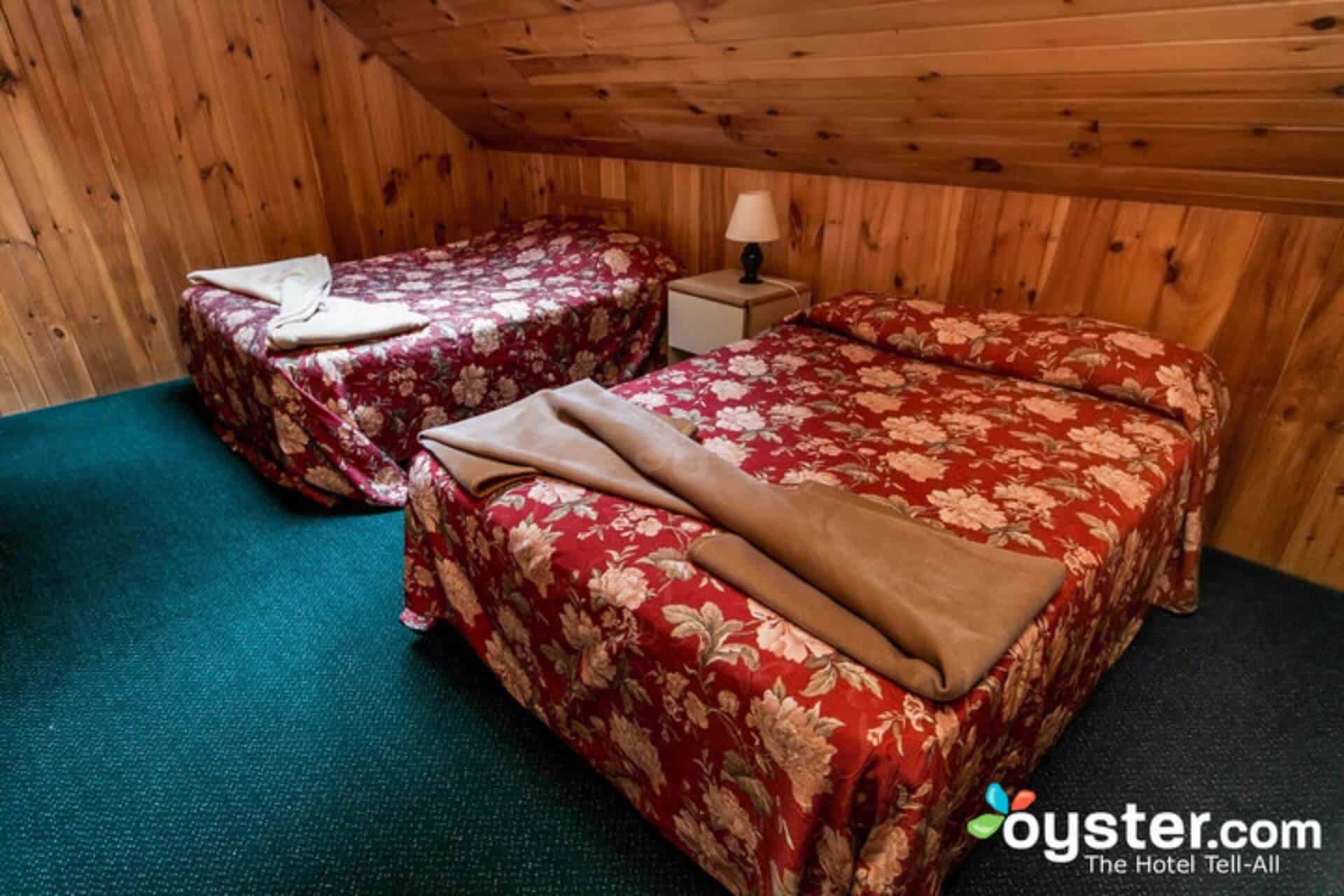 A cozy attic bedroom with sloped wood-paneled walls and a green carpet, featuring two beds with floral bedspreads and a shared nightstand.
