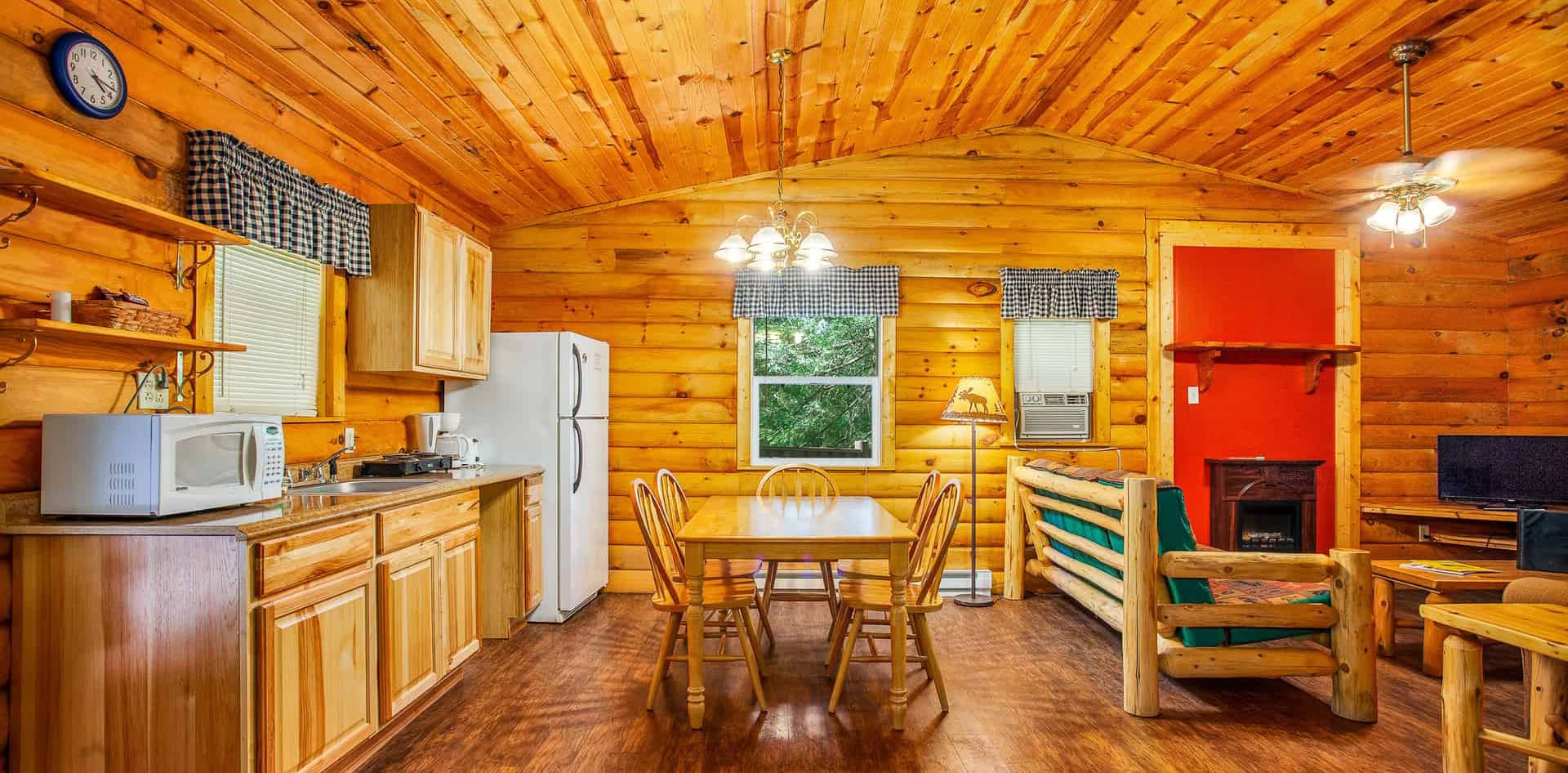 A cozy cabin interior featuring wooden walls, a kitchen area, dining table, and a living space with a sofa and TV.